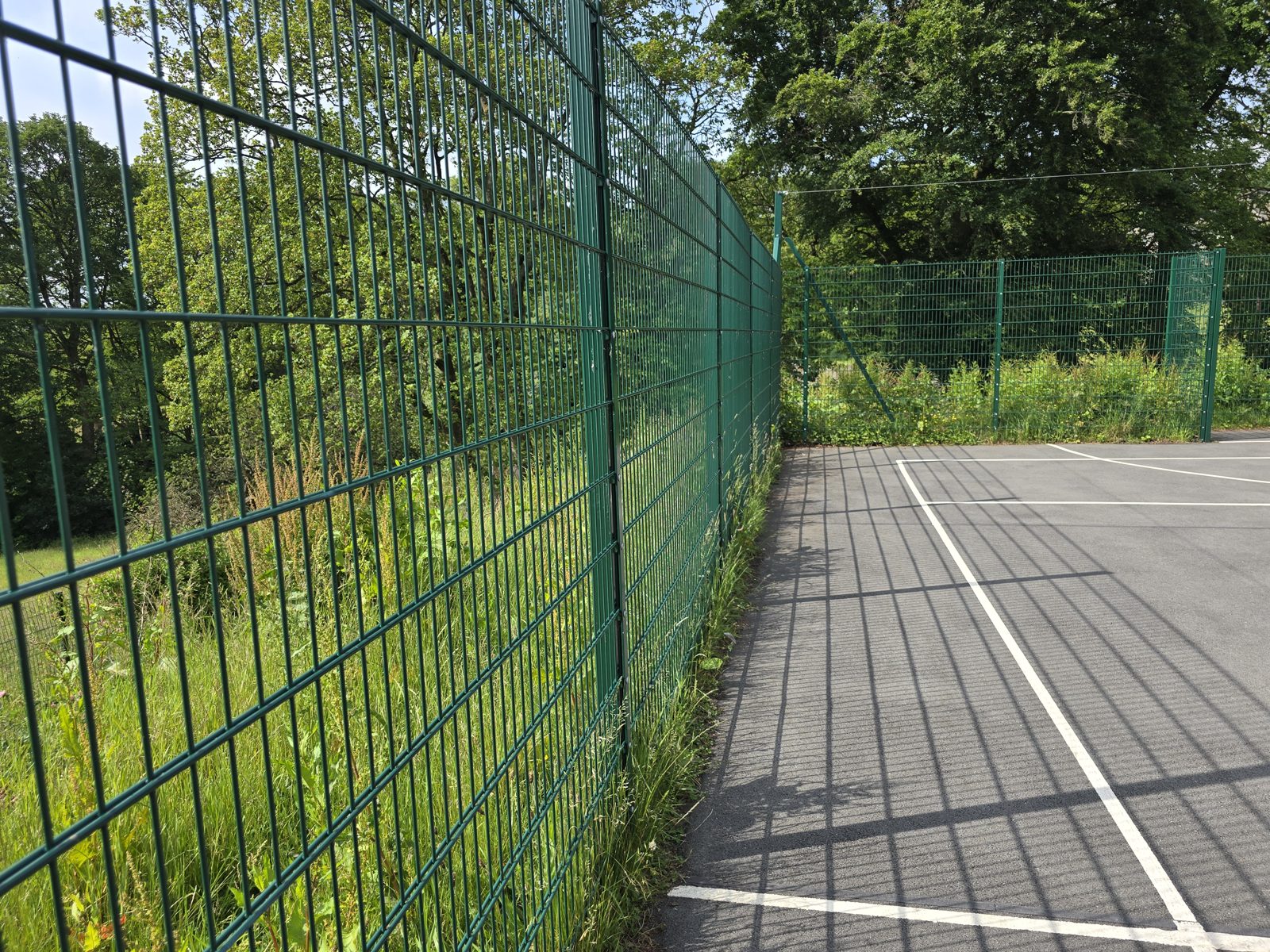 Tall mesh fence beside a marked tarmac games court, top line visibly uneven along the run